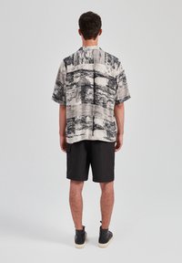 Mężczyzna stojący tyłem, ubrany w koszulę z krótkim rękawem w czarno-białe wzory, czarne szorty i czarne wysokie trampki.