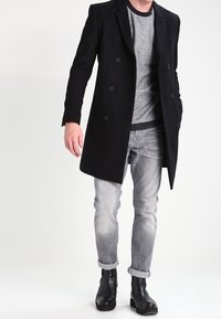 Manteau noir à double boutonnage sur un pull gris, associé à un jean gris clair et des bottines noires, affichant une silhouette élégante et moderne.
