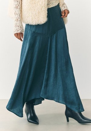 Gonna fluida color teal con finitura testurizzata, caratterizzata da un orlo asimmetrico. Abbinata a stivali neri con punta a livello della caviglia. Sovrapposta a un gilet di pelliccia crema.