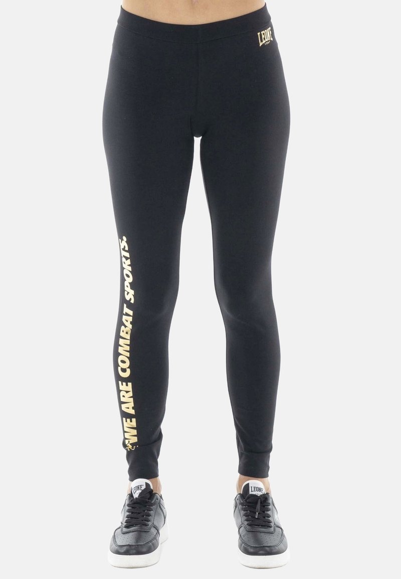 Femme portant un legging noir avec le texte « WE ARE COMBAT SPORTS » sur la jambe gauche et des baskets noires à semelles blanches.