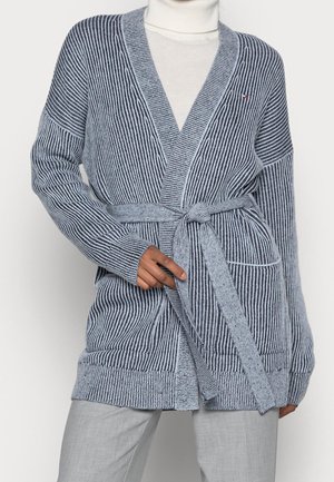 Szary i granatowy prążkowany sweter na guziki z paskiem zawiązanym w talii, noszony na białym golfie i zestawiony z jasnoszarymi spodniami.