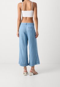 Žena v bledě modrých širokých zkrácených džínách, s bílými sandály na blokovém podpatku a bílém zkráceném topu, stojící před jednoduchým pozadím.