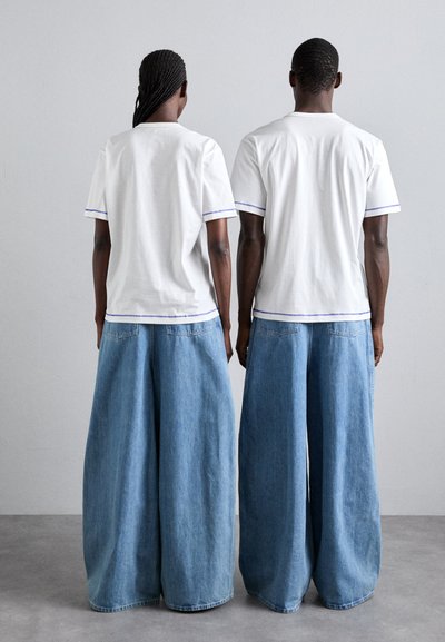 Λευκά μπλουζάκια με μοβ λεπτομέρειες συνδυασμένα με φαρδιά τζιν παντελόνια σε ανοιχτό μπλε χρώμα. Το ύφασμα φαίνεται μαλακό, με χαλαρή εφαρμογή και casual στυλ.