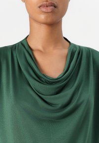 Top verde drappeggiato realizzato in tessuto liscio, con scollatura a grembiule e vestibilità rilassata. Una trama sottile arricchisce il design complessivo.