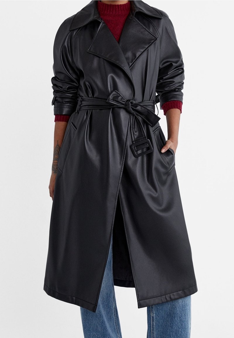 Trench coat in ecopelle nera con vita regolabile, grande colletto e due tasche laterali. Presenta maniche arrotolate e un design lungo fino al ginocchio.