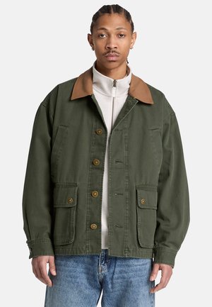 Junger Mann trägt eine olivgrüne Jacke mit braunem Kragen und Knopfdetails, darüber ein weißes Zip-up-Oberteil und blaue Jeans, steht vor weißem Hintergrund.