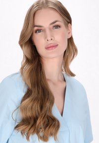 Hellblaue Bluse mit V-Ausschnitt und weicher Textur; das Modell hat lange, wellige blonde Haare und trägt ein natürliches Make-up, während es vor einem weißen Hintergrund steht.