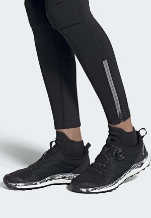 Nohy v černých sportovních legínách se zipy po stranách a černých teniskách Adidas Terrex s bílými vzorovanými podrážkami a šněrováním BOA.