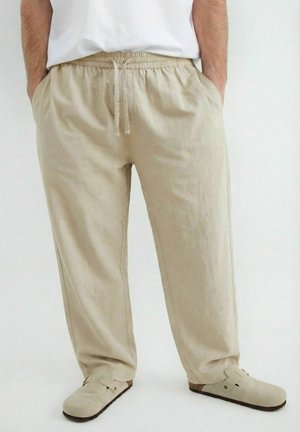 Mężczyzna noszący beżowe luźne spodnie z elastyczną talią i sznurkiem, ręce w kieszeniach, w połączeniu z beżowymi butami wsuwanymi.