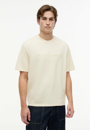 Ung mand iført en cremefarvet Tommy Jeans T-shirt og mørkeblå jeans, stående mod en ensfarvet hvid baggrund.