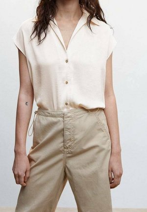 Frau trägt eine cremefarbene, kurzärmelige Bluse mit Knöpfen, die in eine hoch taillierte beige Hose gesteckt ist, vor einem einfarbigen Hintergrund.
