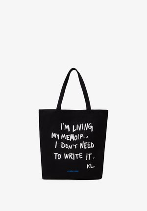 Bolsa tote negra con dos asas, texto escrito a mano en blanco que dice "Estoy viviendo mi memoria. No necesito escribirla." y pequeño hashtag azul #KARLCORE.