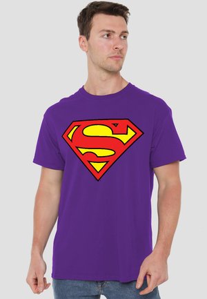 Man wearing purple t-shirt with large red and yellow Superman logo, looking to his right against plain background.