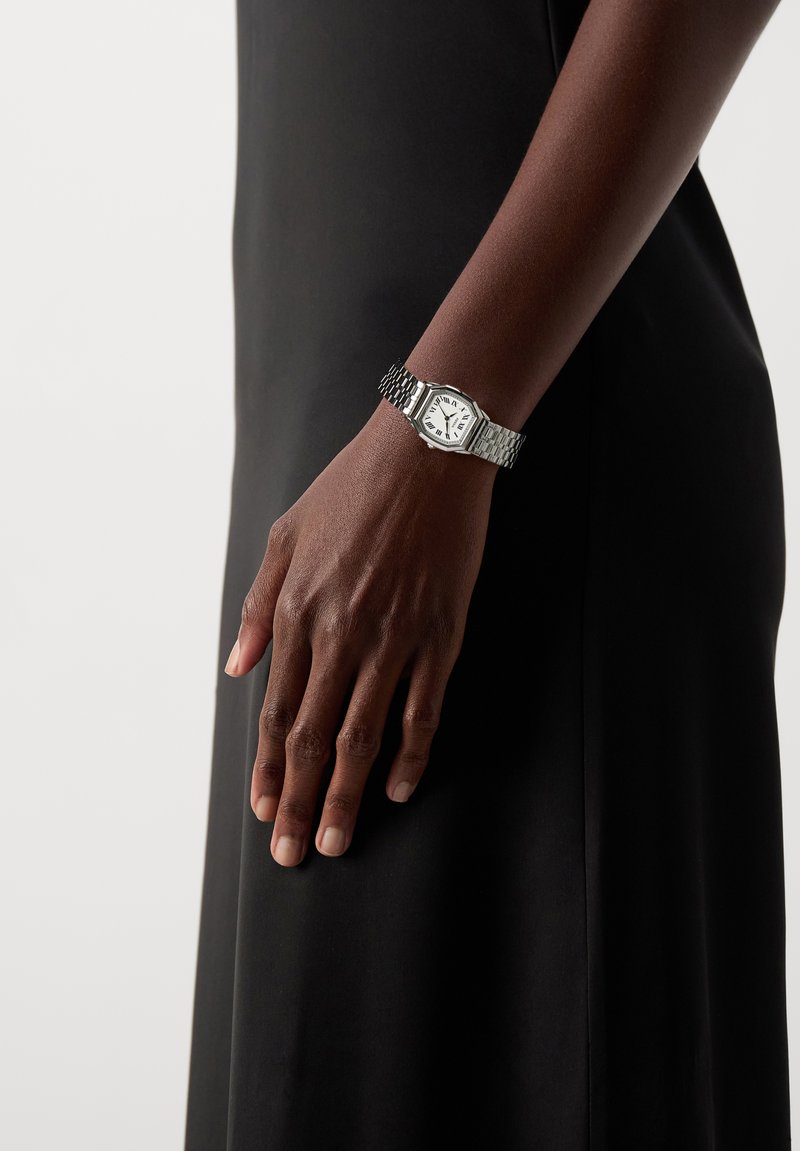 Σκούρο δέρμα χέρι που φορά ένα ασημένιο μεταλλικό ρολόι με λευκό καντράν και ρωμαϊκούς αριθμούς, που ακουμπάει σε μαύρο φόρεμα.
