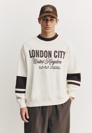 Cremefarbener Sweatshirt mit maronenfarbenen Akzenten, versehen mit fettem Text: "LONDON CITY United Kingdom 207-203 STUDIOS." Kombiniert mit braunen karierten Hosen.