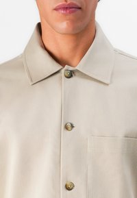 Man wearing beige button-up shirt with collar and chest pocket, viewed from shoulders to mouth against white background.