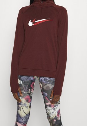 Personne portant un pull bordeaux à manches longues avec un logo swoosh blanc et rouge et des leggings à motifs floraux colorés, debout sur un fond blanc.