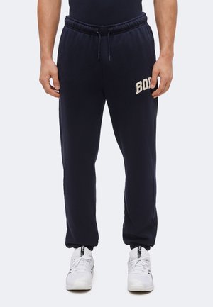 Homme portant un pantalon de jogging noir avec le texte "BOSS" sur la cuisse gauche et des baskets blanches sur un fond clair uni.