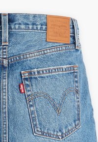 Jeans in denim blu con etichetta in pelle marrone sulla cintura. Presentano una tasca posteriore con cuciture distintive e un piccolo etichetta rossa del marchio.