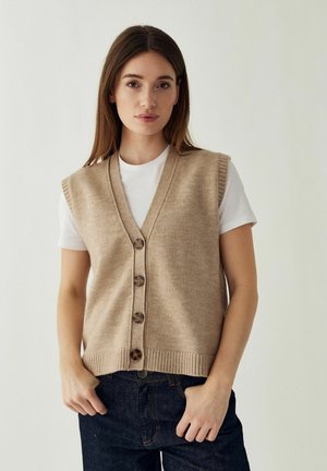 Donna con capelli castani lunghi che indossa un gilet lavorato a maglia beige senza maniche con bottoni sopra una camicia bianca a maniche corte e jeans scuri, in piedi davanti a uno sfondo uniforme.