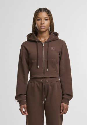 Hnedá skrátená mikina s kapucňou z mäkkej tkaniny, vybavená predným zipsom, dvoma vreckami a kapucňou so šnúrkou a rebrovanými manžetami.