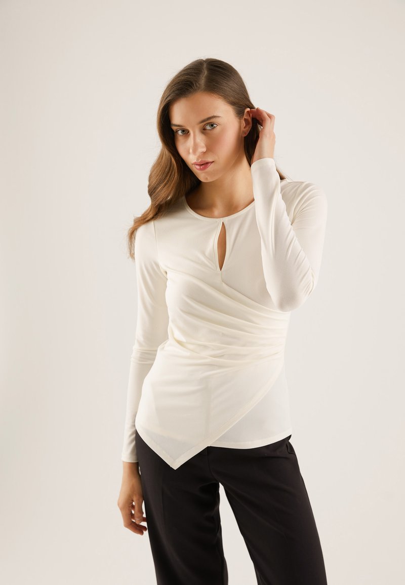 Long-sleeve white top with a keyhole neck, fitted body, asymmetric hem, and gathered texture, paired with black pants.