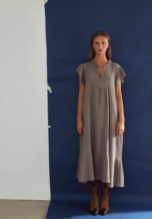 Kvinde står foran en mørkeblå baggrund iført en løs, grå midi-kjole med korte ærmer og brune cowboystøvler.