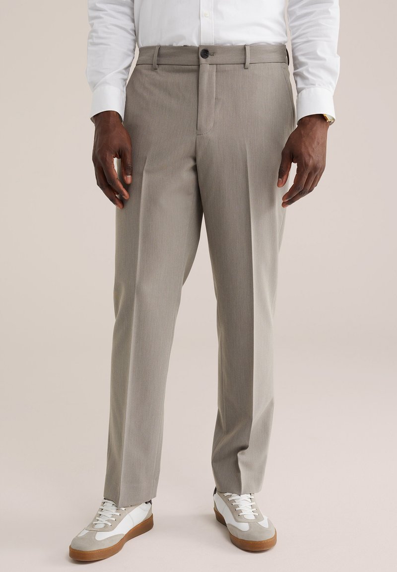 Homme portant un pantalon beige, une chemise blanche et des baskets blanches, debout devant un fond uni, les mains détendues le long du corps.