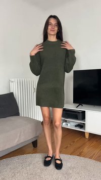 Vestito verde a maglia con maniche lunghe, collo rotondo e vestibilità ampia, abbinato a ballerine nere. Sullo sfondo si vedono un termosifone e una televisione.