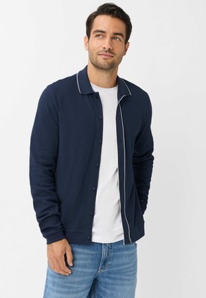 Mand iført en navyblå knap-cardigan over en hvid t-shirt og blå jeans, stående med den ene hånd i lommen og kigger til siden.
