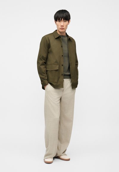 Άνδρας με χακί πράσινο σακάκι με κουμπιά πάνω από πράσινο πουλόβερ, μπεζ παντελόνια με φαρδιά πόδια και λευκά παπούτσια, στέκεται με τα χέρια στις τσέπες.