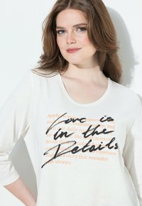 Donna con pelle chiara e capelli castani che indossa una camicia bianca con la scritta "L'amore è nei dettagli" sovrastata da un testo arancione.