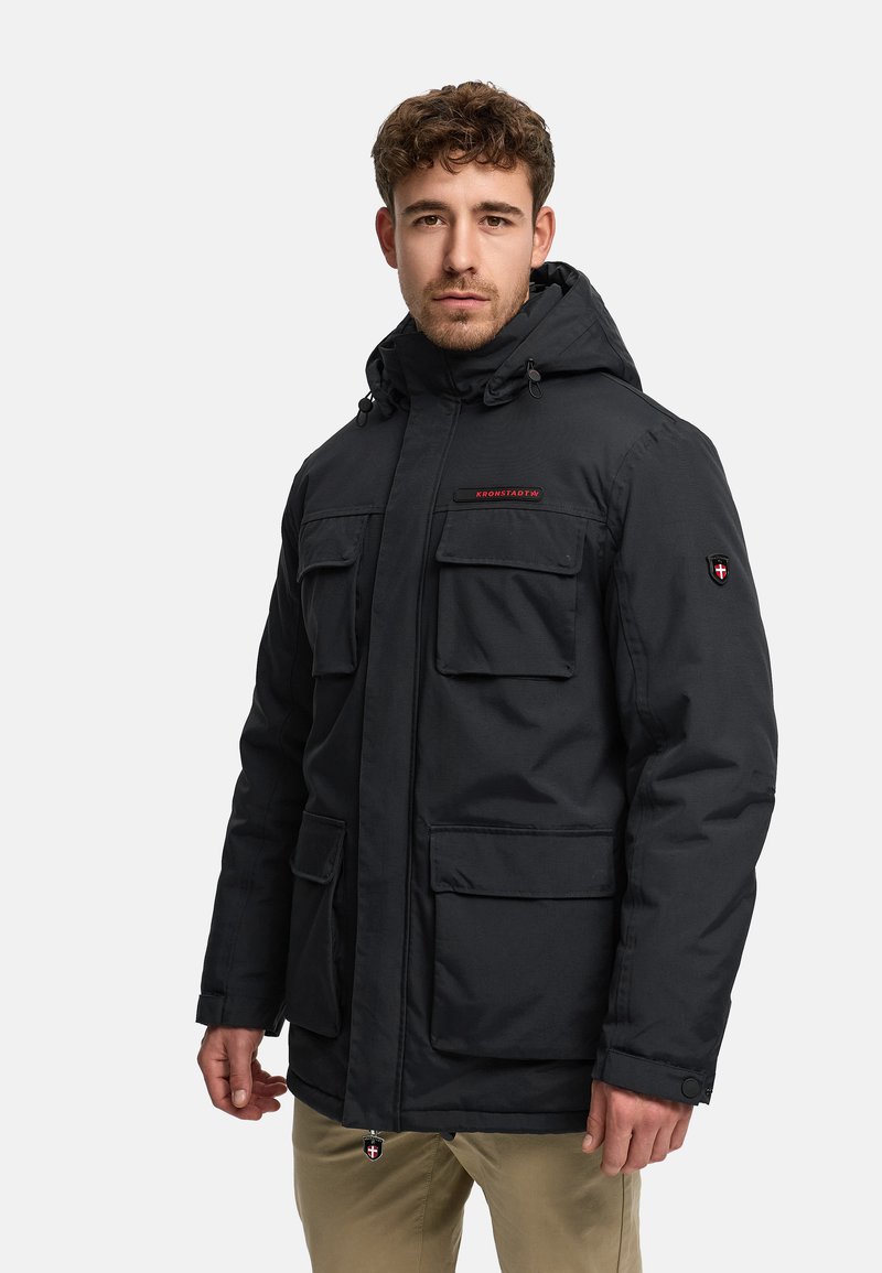 Man draagt een zwarte gewatteerde jas met klepzakken en beige broek, staand tegen een effen witte achtergrond.