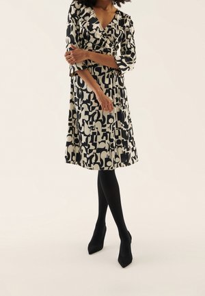 Femme portant une robe portefeuille à motif abstrait noir et blanc avec des manches trois-quarts, associée à un collant noir et des talons noirs.