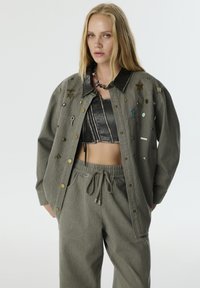 Oversized grijze jas met verschillende decoratieve spelden, gecombineerd met een zwart cropped top en bijpassende grijze broek met een elastische tailleband.