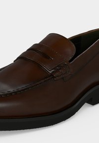 Mocassin penny en cuir marron avec surpiqûres moc toe et semelle en caoutchouc noir sur fond blanc.