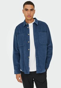 Marineblå fløjlsskjorte med knapfront, to brystlommer og lange ærmer, lagd over en hvid t-shirt. Enkel design med struktureret stof.