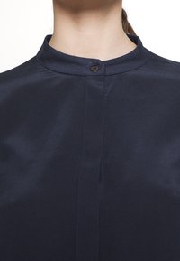 Granatowa bluzka z kołnierzykiem mandarynkowym, gładka faktura, zapięcie na jeden guzik oraz wyraźny szew biegnący wzdłuż przodu.