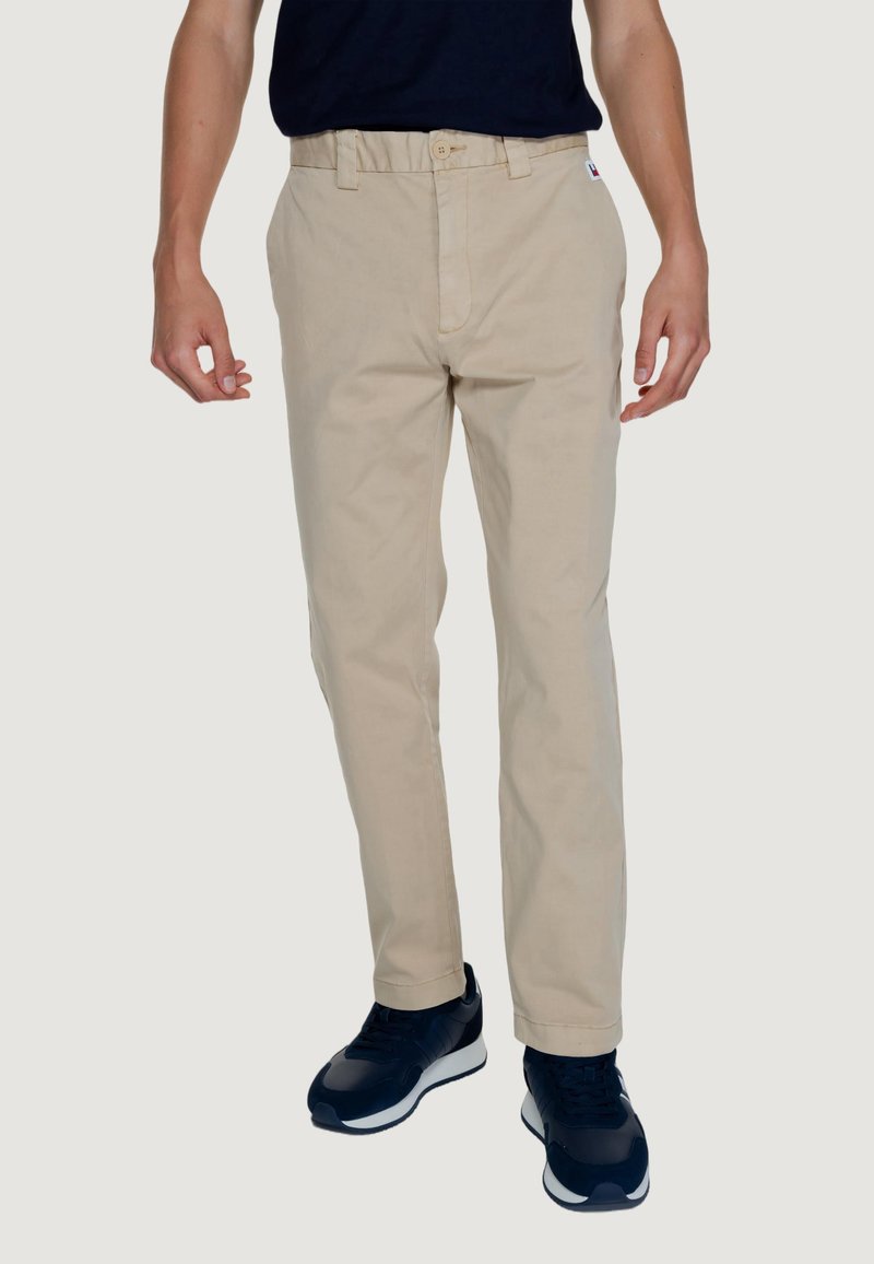 Pantaloni beige in cotone dal taglio dritto, con chiusura frontale a bottoni e tasche laterali, indossati con sneakers blu navy.