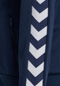 Giacca blu navy con zip, caratterizzata da un motivo a zigzag bianco sul lato. Il tessuto presenta una finitura testurizzata, che ne evidenzia la durevolezza e il comfort.