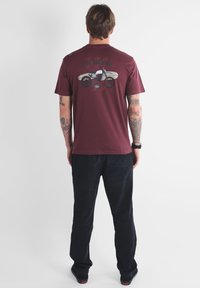 Homme avec des tatouages portant un t-shirt bordeaux avec un graphisme de moto et de planche de surf, un pantalon noir et des chaussures noires, se tenant de dos.