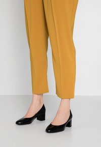 Escarpins en cuir verni noir à talons carrés et à bout rond, associés à un pantalon droit en jaune moutarde.
