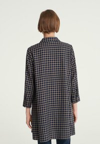 Mörkblå rutmönstrad tunika med ljusbeige rutmönster, långa ärmar och rak skärning. Har en krage och knäppta ärmmuddar.