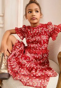 Rotes Kleid mit kurzen Puffärmeln, das ein Herzmuster in Pink und Gold aufweist. Mit einem Rüschensaum und einem geschichteten Design. Getragen mit metallischen goldenen Stiefeln.