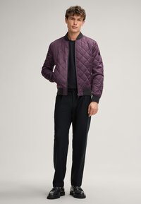 Veste bomber matelassée violette avec des poignets et un col côtelés noirs, associée à un pantalon noir et des chaussures en cuir, sur un fond neutre.