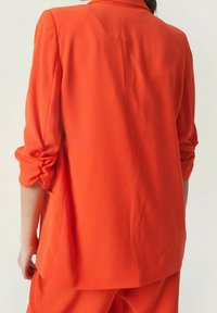 Jasnopomarańczowa marynarka z podwiniętymi rękawami, gładką fakturą, pojedynczym wentylem z tyłu i prostymi liniami. Materiał wydaje się lekki i zwiewny.