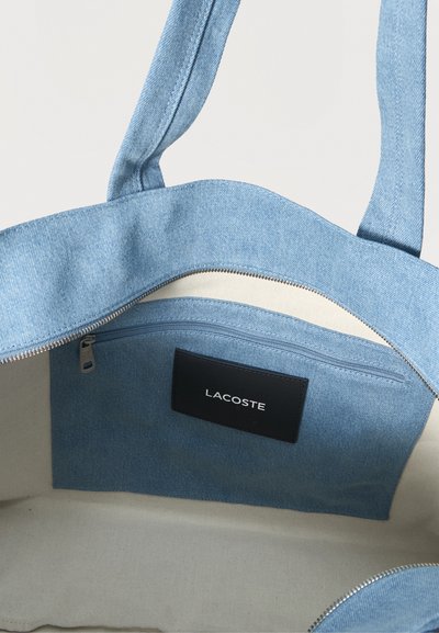 Gros plan sur un sac cabas en denim bleu, ouvert par le haut et doté d’une fermeture éclair, d’une doublure beige et d’une étiquette Lacoste noire.