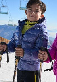 Chaqueta acolchada morada con textura brillante, cuello amarillo con rayas negras y bastones de esquí en las manos; fondo nevado con telesilla.