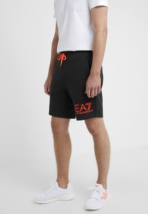 Zwarte shorts met een elastische tailleband, oranje trekkoord en contrasterende logoafdruk op het linkerbeen. Draag je met witte sportschoenen.