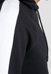 Detailní záběr osoby v černé mikině s bílým panelem na rukávu a černou šňůrkou na kapuci.